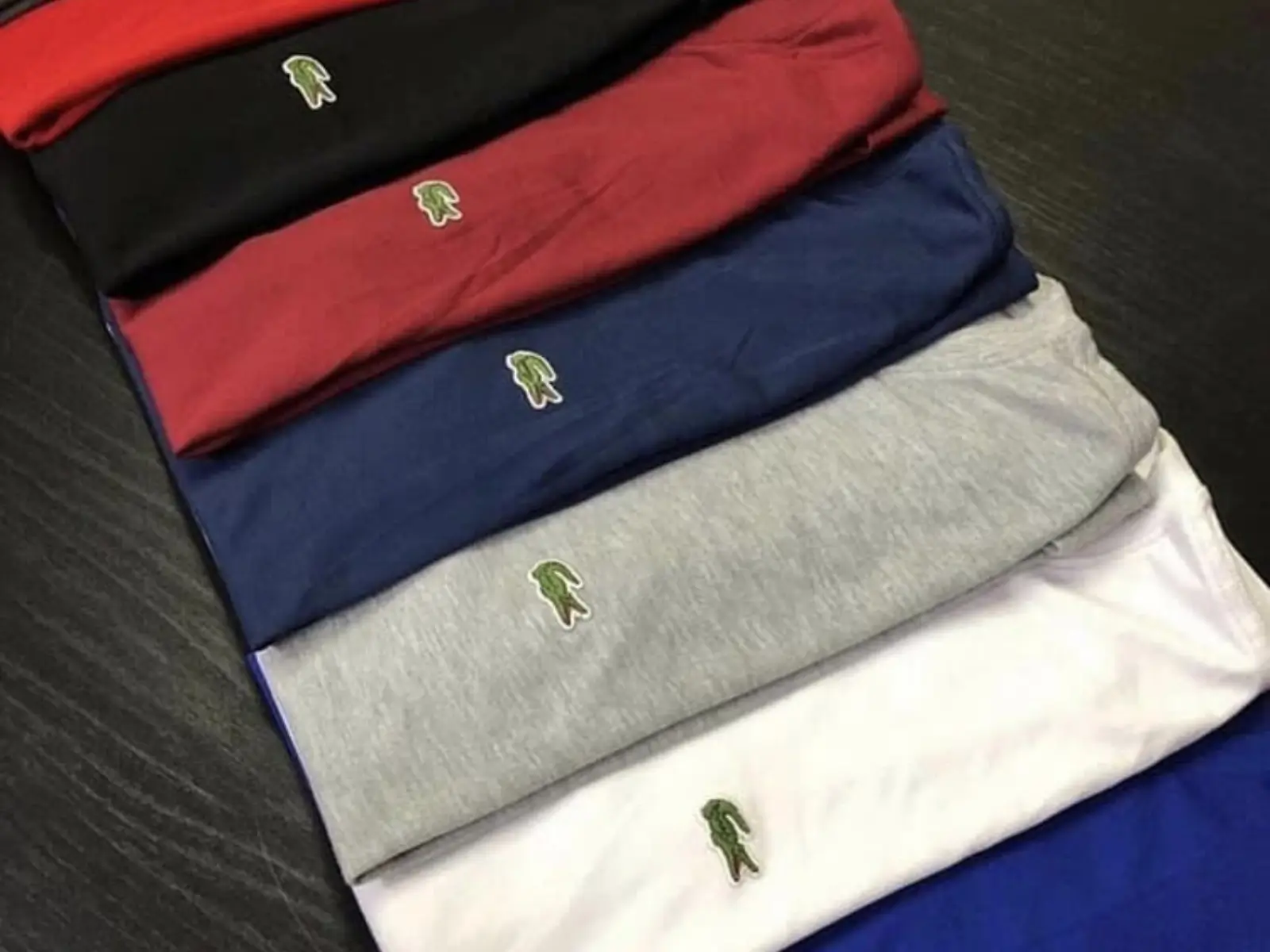 Pilha de camisetas básicas da Lacoste em cores variadas, como preta, vermelha e azul, disponíveis com cupom de desconto. Cupom de Desconto Lacoste