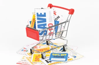 Um carrinho de compras em miniatura cheio de cupons de desconto, com destaque para a palavra "SAVE" (economize). Vários outros cupons estão espalhados ao redor, sobre um fundo branco, simbolizando a economia na primeira compra.