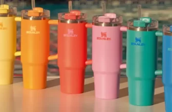 Fileira de copos Stanley Quencher em diversas cores vibrantes, como amarelo, laranja, rosa e azul, posicionados ao lado de uma piscina, representando a coleção de verão da marca.