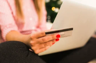 Close-up da mão de uma mulher com unhas vermelhas segurando um cartão de crédito em frente a um notebook, no momento de finalizar uma compra online com cupom de desconto.