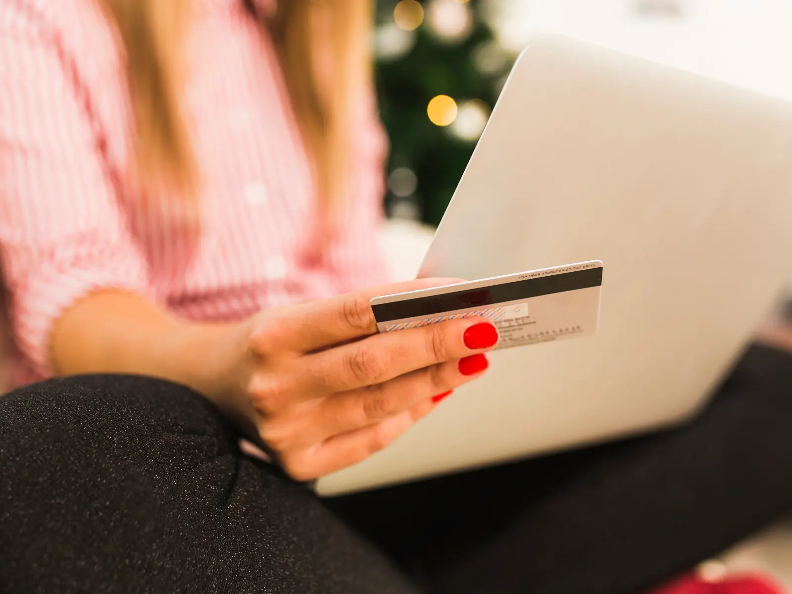 Close-up da mão de uma mulher com unhas vermelhas segurando um cartão de crédito em frente a um notebook, no momento de finalizar uma compra online com cupom de desconto.
